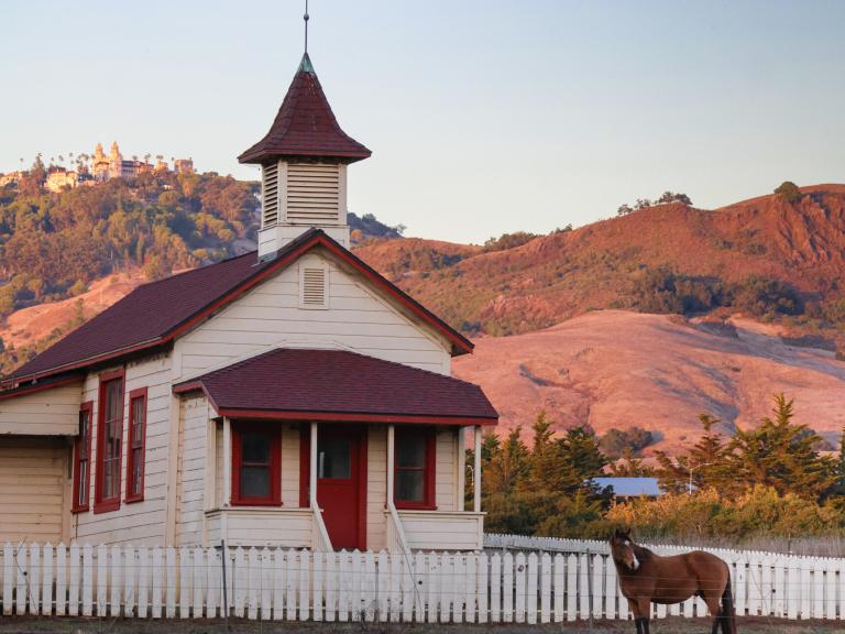 A historic white schoolhouse with red trim sits below rolling hills at Hearst San Simeon State Park and Hearst Castle in the background.