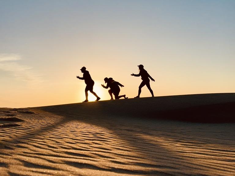 Three silhouetted people walking across the dunes at sunset, their shadows stretching over the sand at Oceano Dunes State Park.