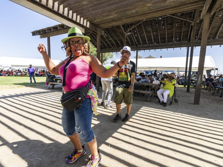 A person in a bright pink top and green hat dances energetically under a wooden pavilion at an outdoor gathering.