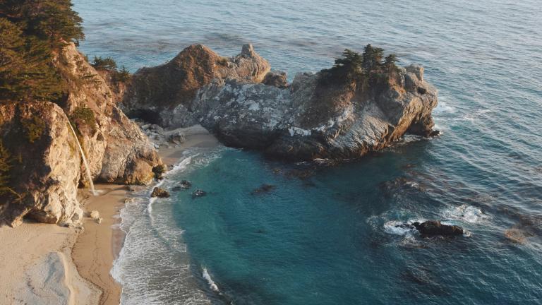 aerial view of ocean beach cove with gentle waves and deep blue waters fading into a pastel horizon softly lit by the afternoon sun